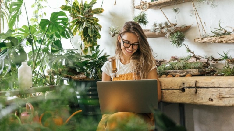 Propietaria de una tienda de plantas que usa gafas y el cabello suelto y que está mirando una computadora portátil.