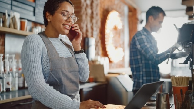 Coffee shop owner in apron and glasses working on a laptop and having a conversation on a mobile phone.