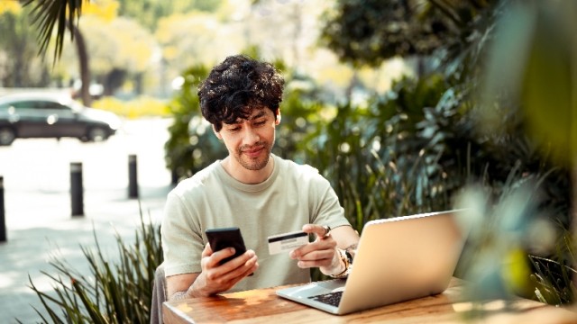 A person sitting outside at a table, checking their computer and phone with a credit card in-hand.