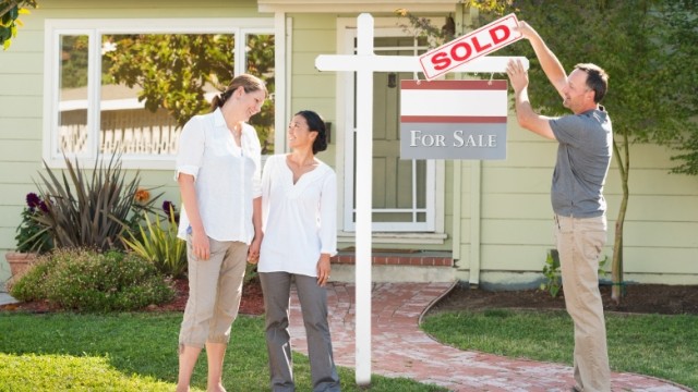 Partners with real estate agent in front of a home with a âSOLDâ sign.