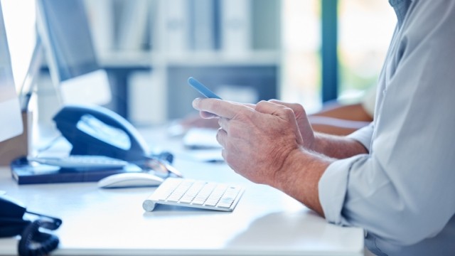 Personâs hands at a desk looking at a mobile phone.