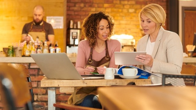 Two women in a coffeeshop talking with a laptop and tablet.