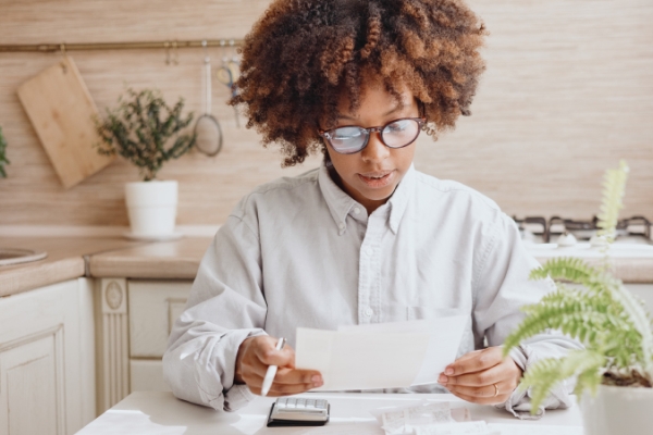 Woman with glasses reviewing finances with calculator at the kitchen table. 