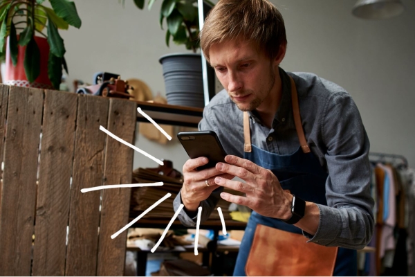 Man in shop apron sits in a chair, checking his phone. 