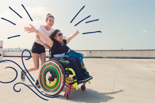 Teenage girl pushing friend in wheelchair with sunglasses on along the sunny beach sidewalk
