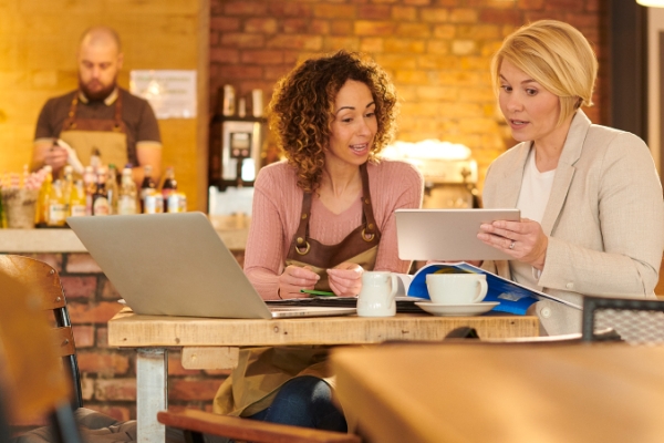 Two women in a coffeeshop talking with a laptop and tablet.